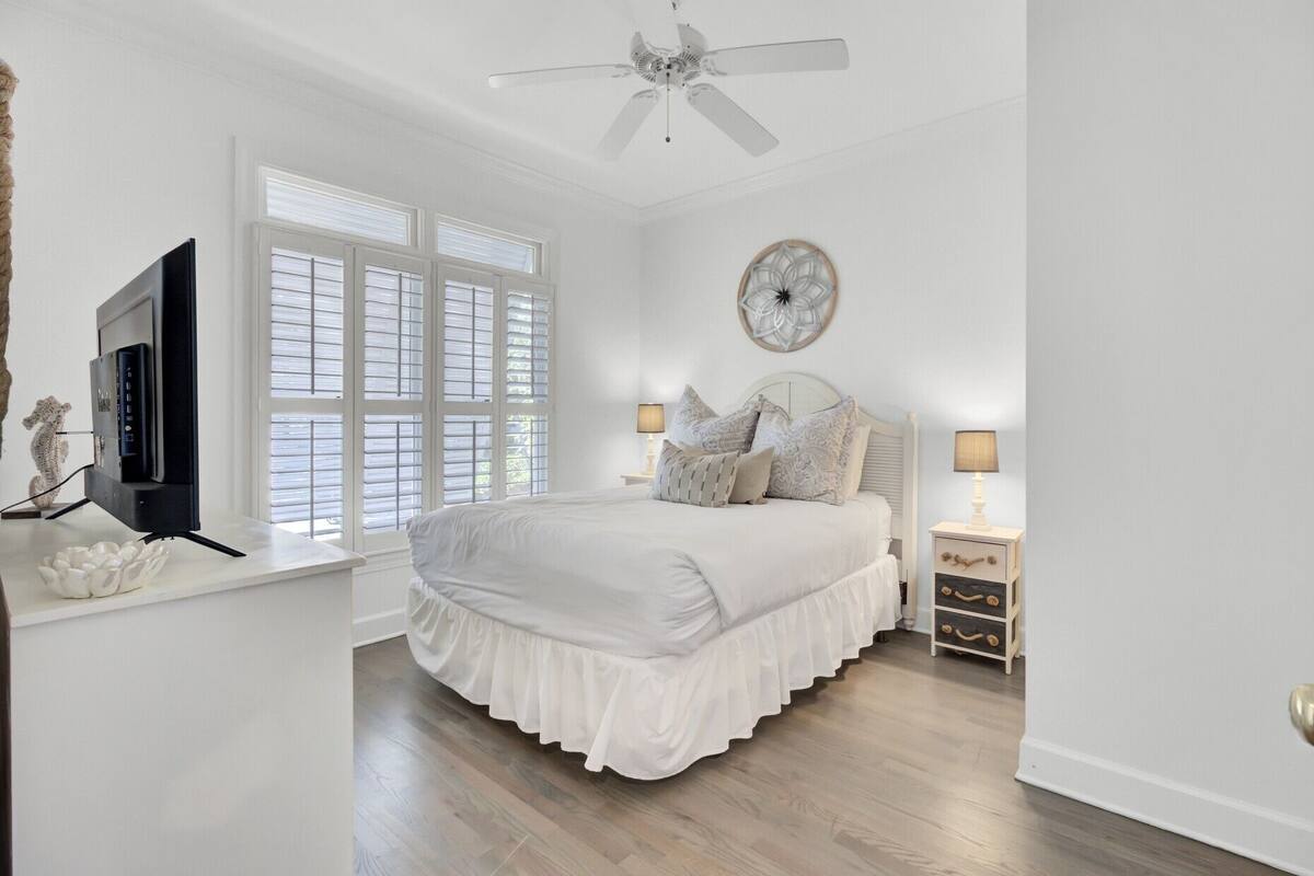 A bedroom with a bed, a ceiling fan, a TV, a window with blinds, a wall clock, and bedside tables with lamps.