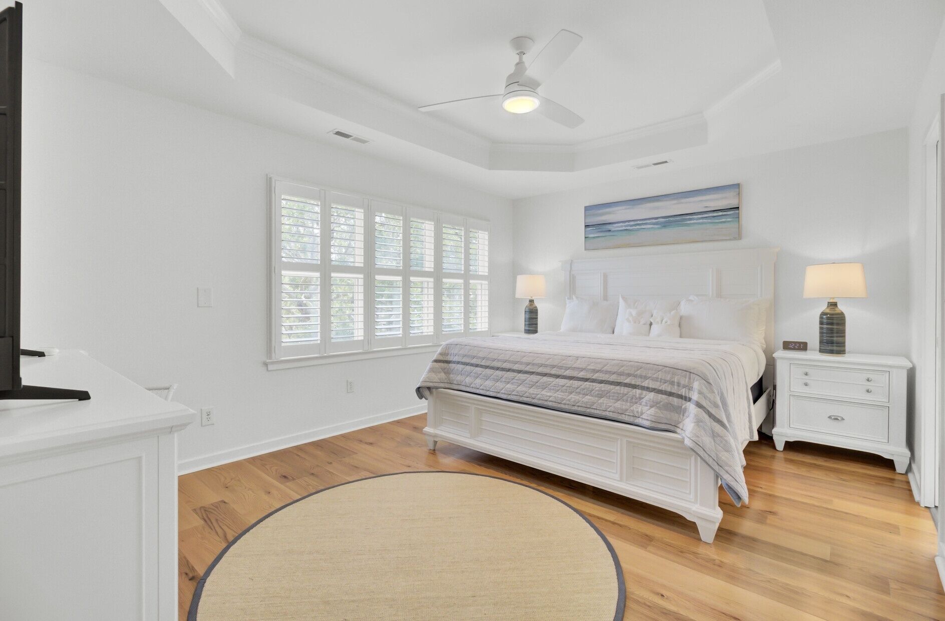 A bedroom with a bed, bedside tables, a ceiling fan, a window with blinds, and a painting on the wall.