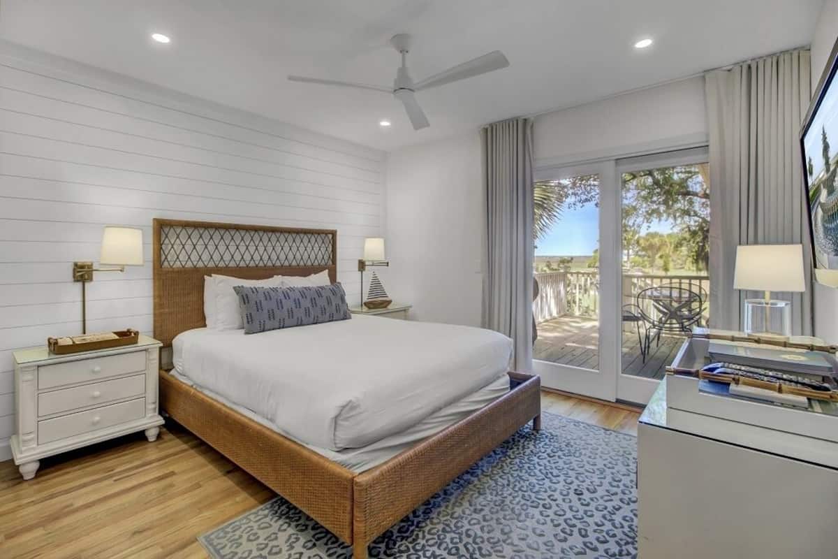 A bedroom with a large bed, a ceiling fan, a nightstand, a desk, and a view of the outdoors.