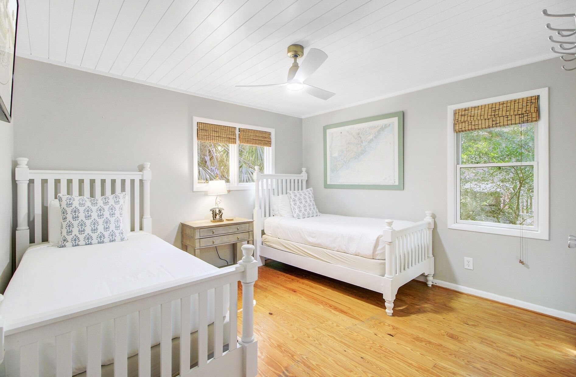 A bedroom with two white beds, a ceiling fan, a nightstand, and a window with a view of greenery.