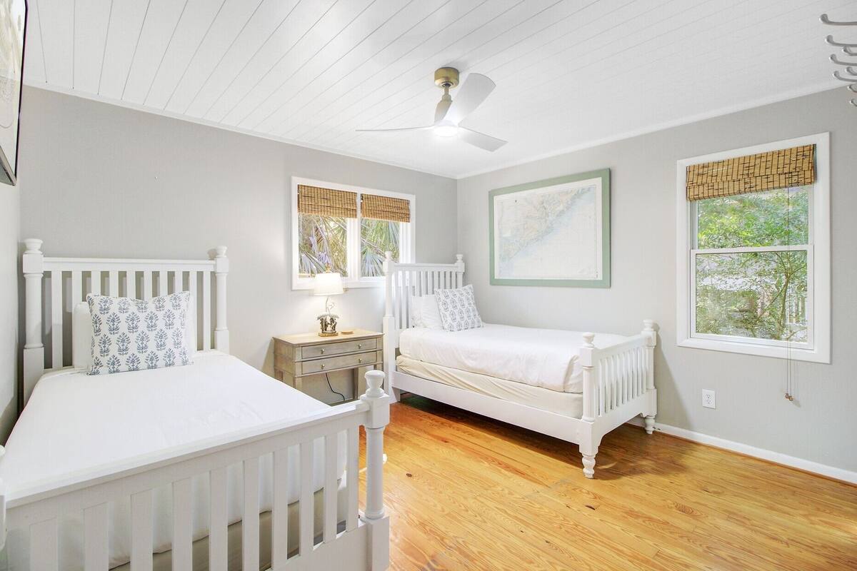A bedroom with two white beds, a ceiling fan, a nightstand, and a window with a view of greenery.