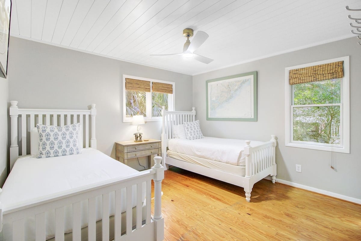 A bedroom with two white beds, a ceiling fan, a nightstand, and a window with a view of greenery.