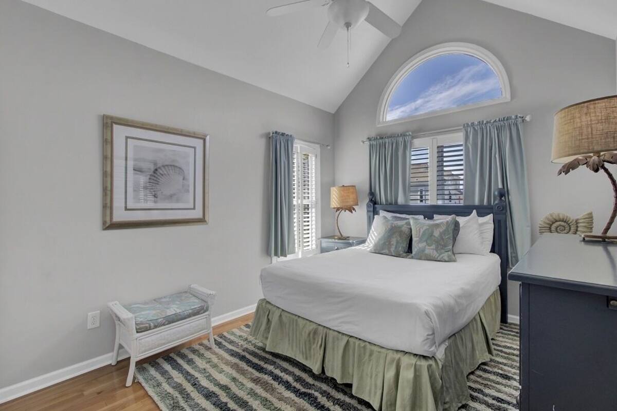 A bedroom with a bed, a bench, a desk, a lamp, a ceiling fan, and a window with curtains.