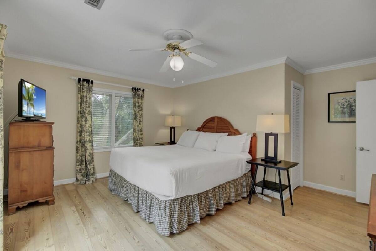 A bedroom with a bed, a ceiling fan, a nightstand, a lamp, a dresser, and a window with curtains.