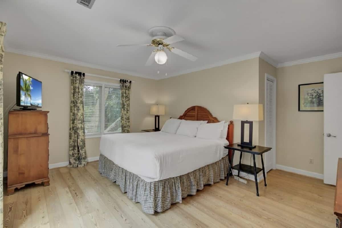 A bedroom with a bed, a ceiling fan, a nightstand, a lamp, a dresser, and a window with curtains.