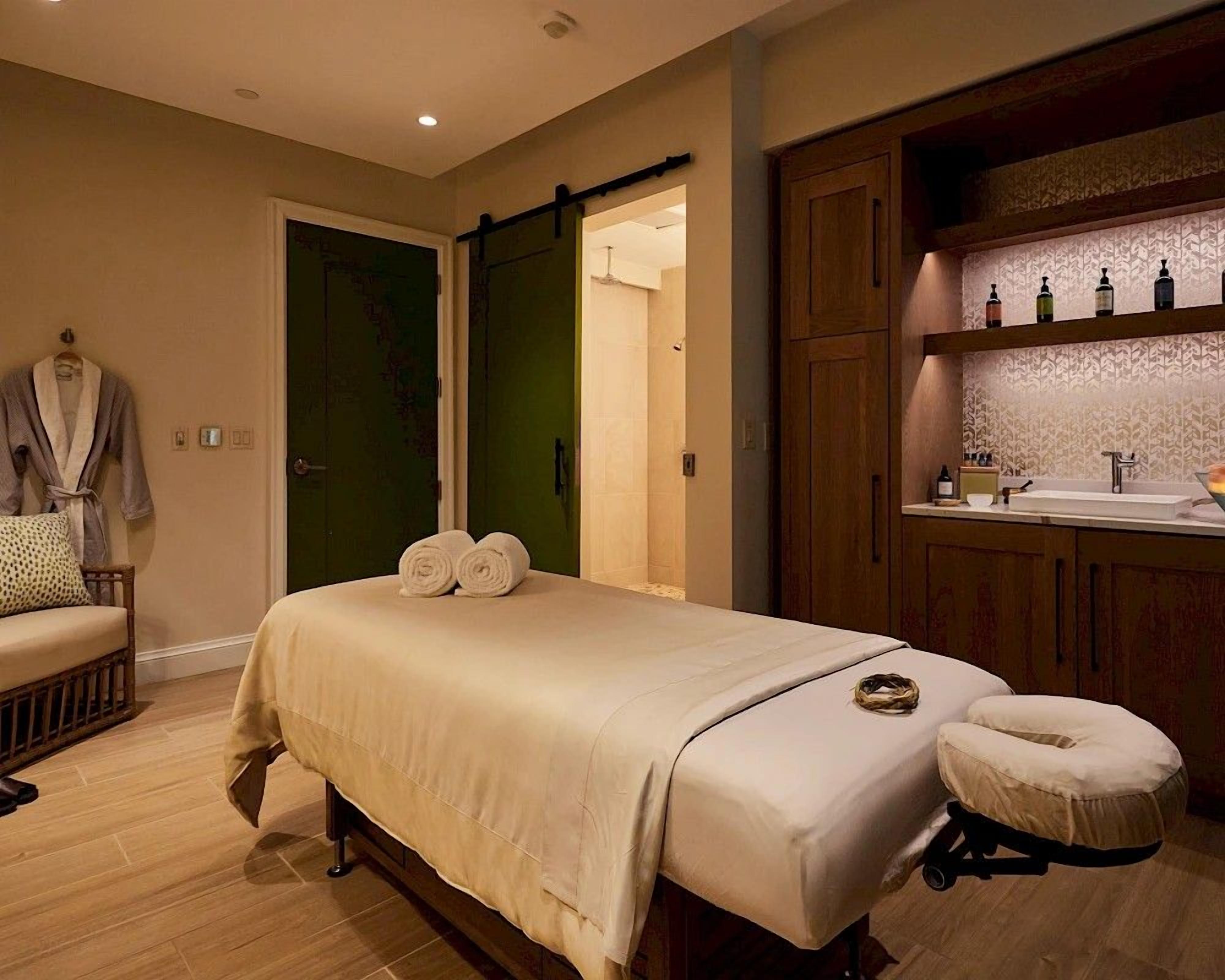 A room with a bed, a massage table, a chair, a green door, and a wooden cabinet with bottles.