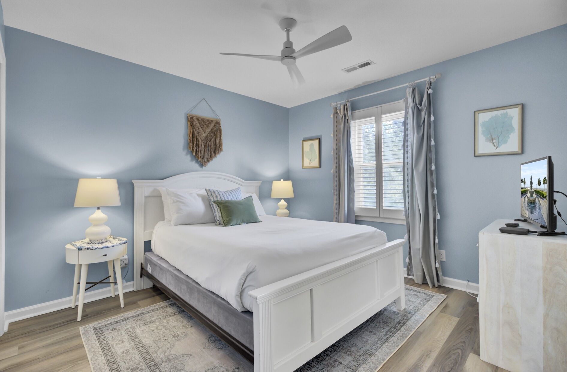 A bedroom with a bed, two bedside tables with lamps, a ceiling fan, a window with curtains, and a television on a stand.