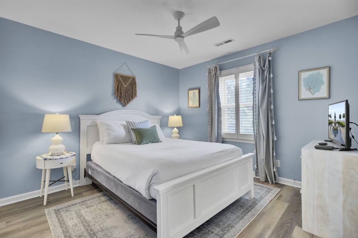 A bedroom with a bed, two bedside tables with lamps, a ceiling fan, a window with curtains, and a television on a stand.