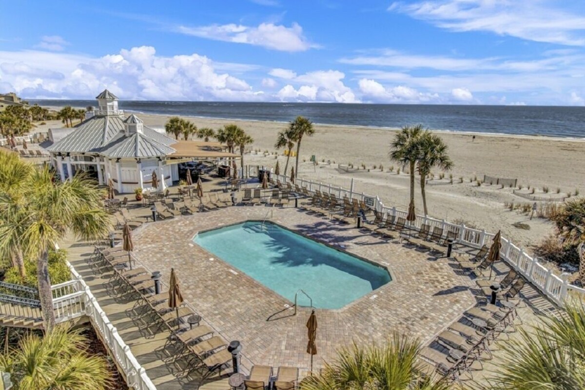 Ein Poolbereich mit Liegestühlen, Sonnenschirmen, einem Pavillon und Blick auf den Strand.