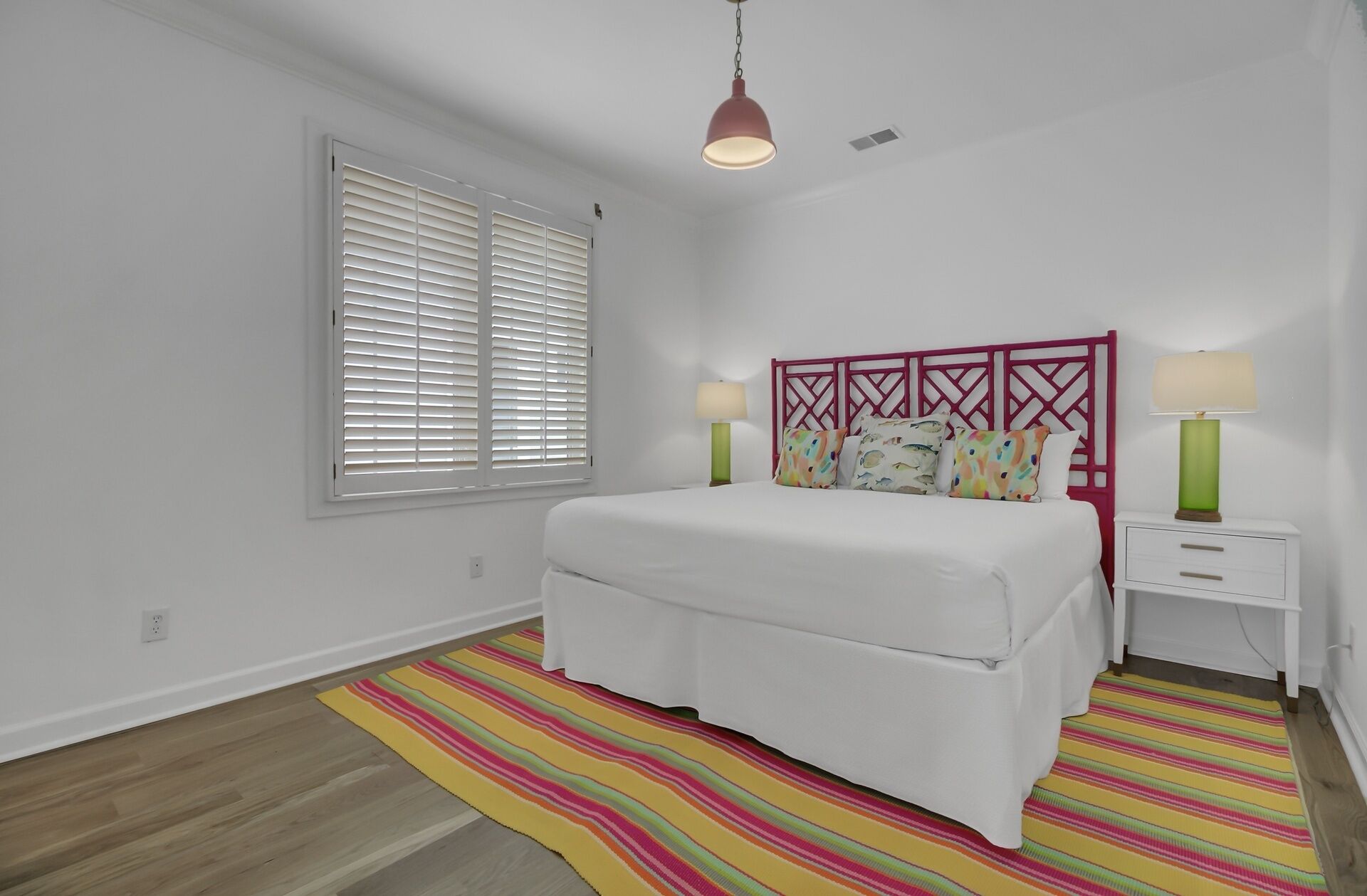 Ein Schlafzimmer mit einem Bett, zwei Nachttischen, einem Fenster mit Jalousien, einem farbenfrohen Streifen Teppich und einer Pendelleuchte.