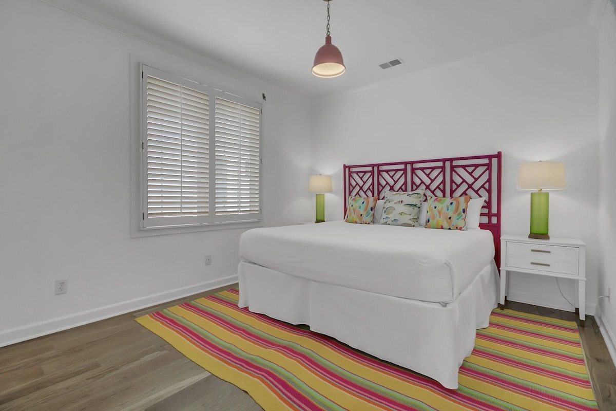 Ein Schlafzimmer mit einem Bett, zwei Nachttischen, einem Fenster mit Jalousien, einem farbenfrohen Streifen Teppich und einer Pendelleuchte.