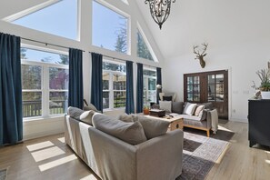 A spacious living room with a large sofa, a coffee table, and a fireplace. There are large windows with curtains, a chandelier, and a deer head decoration on the wall. - The Rail & Pine Spacious Masthope Mountain Retreat (Lackawaxen)