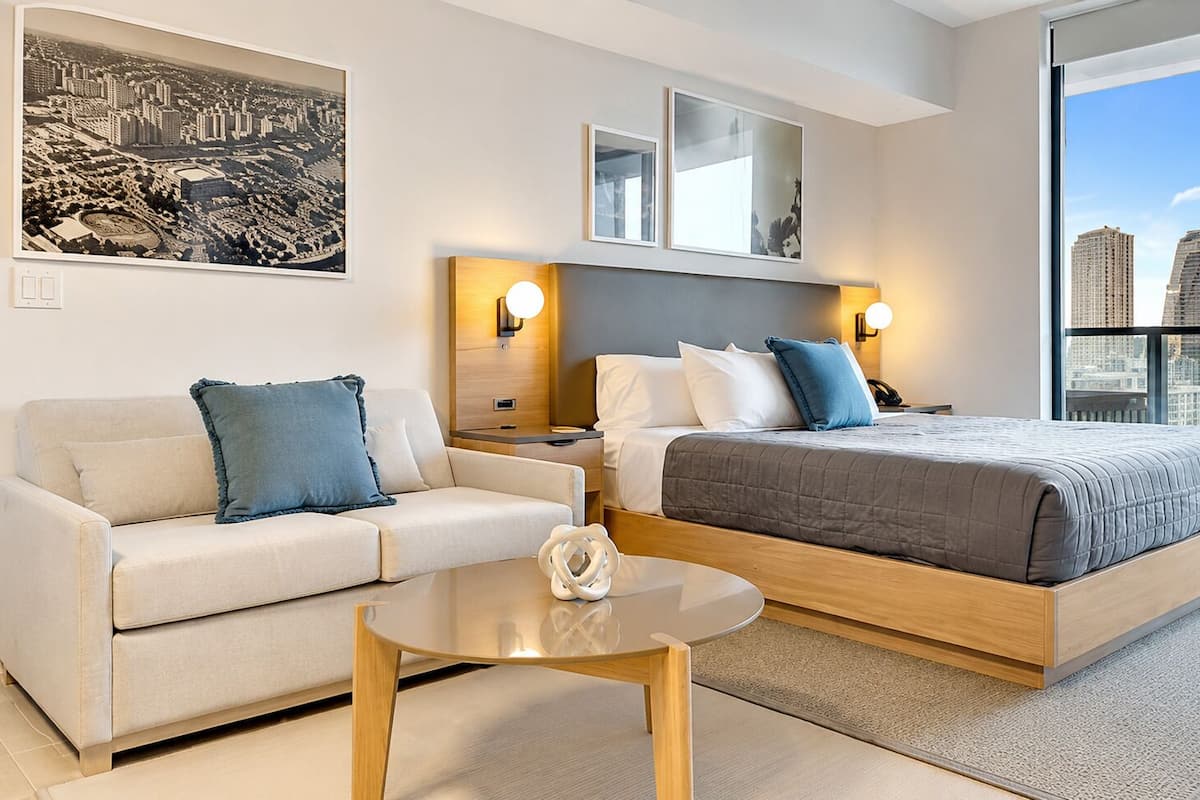 A modern hotel room with a bed, a sofa, a round coffee table, and a large window with a city view.