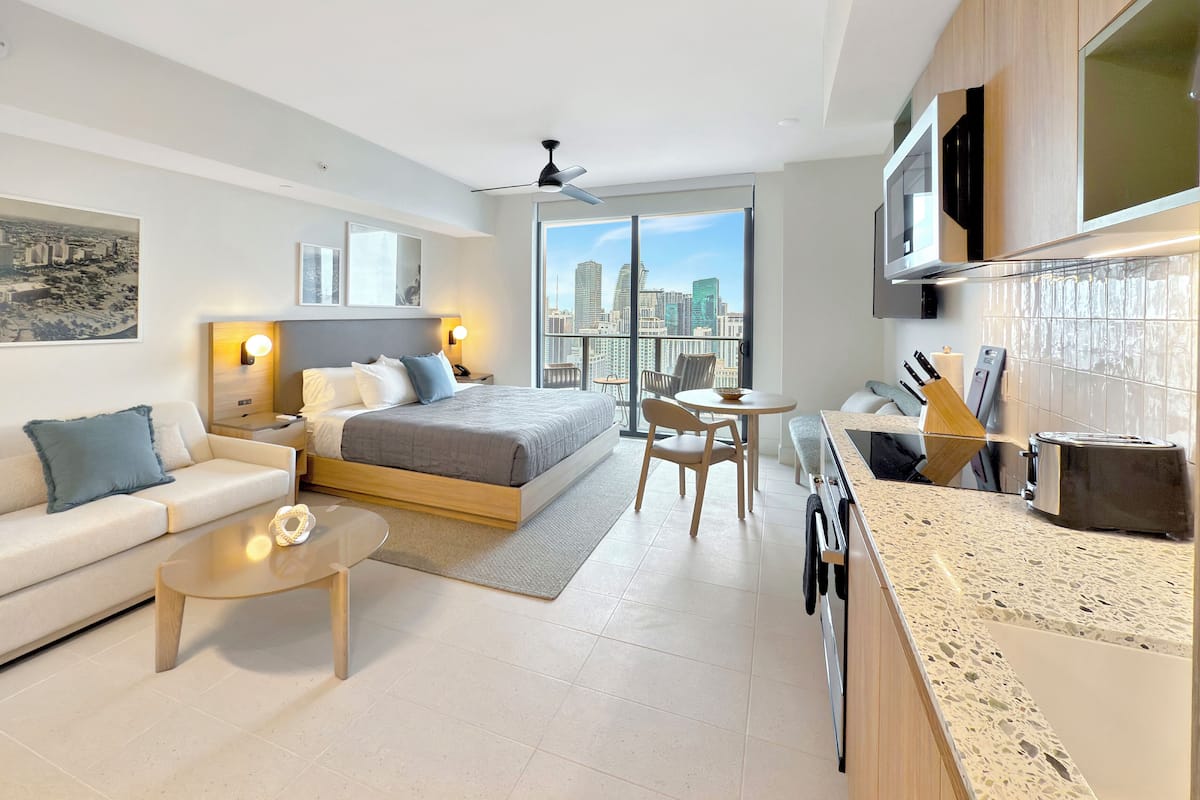 A modern hotel room with a bed, sofa, coffee table, and a kitchen area with a TV and microwave.