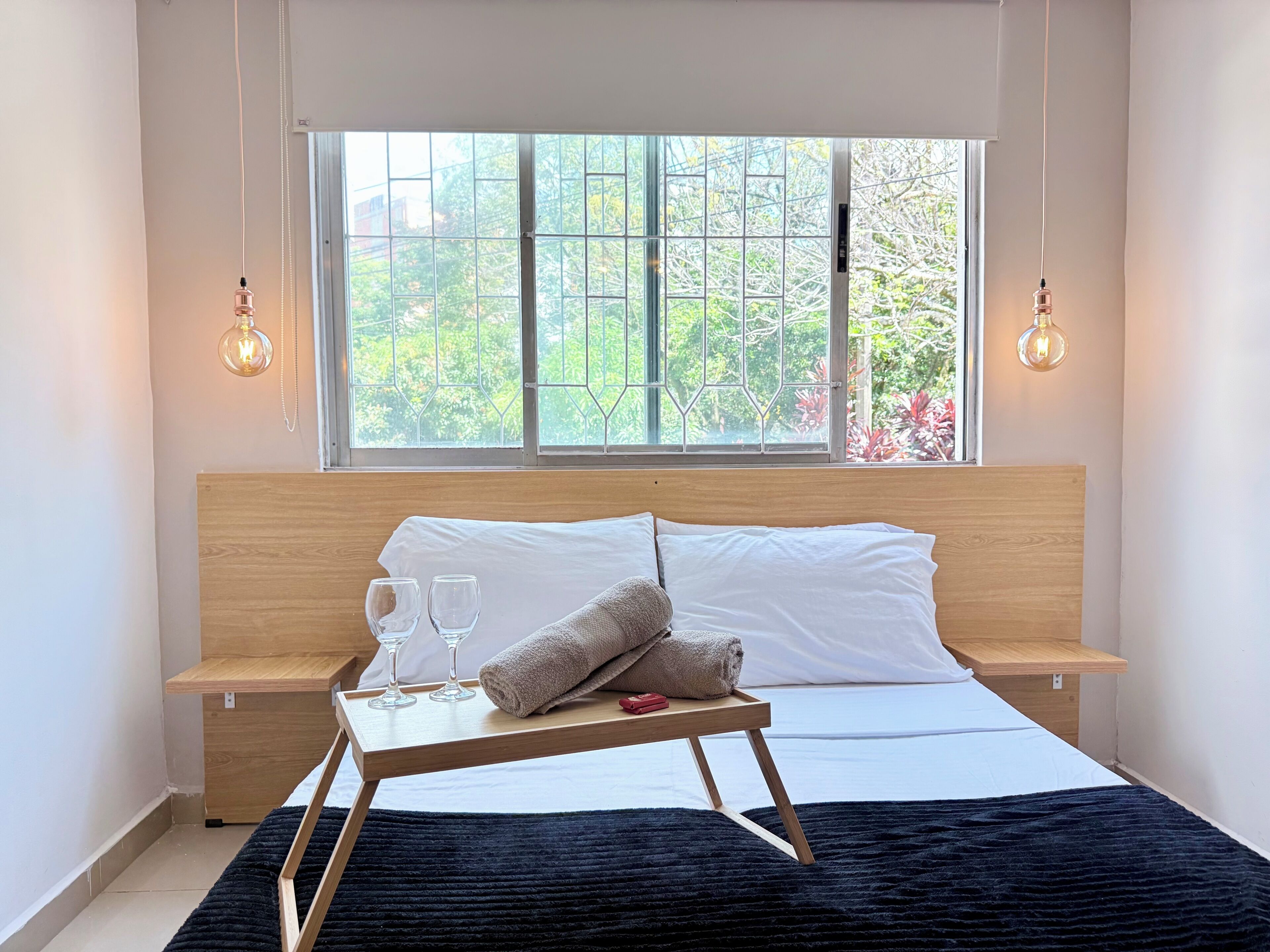 Et soveværelse med sengegavl i træ, et vindue med udsigt til grønne omgivelser, sengeborde med lamper og et bakke med glas og håndklæde.