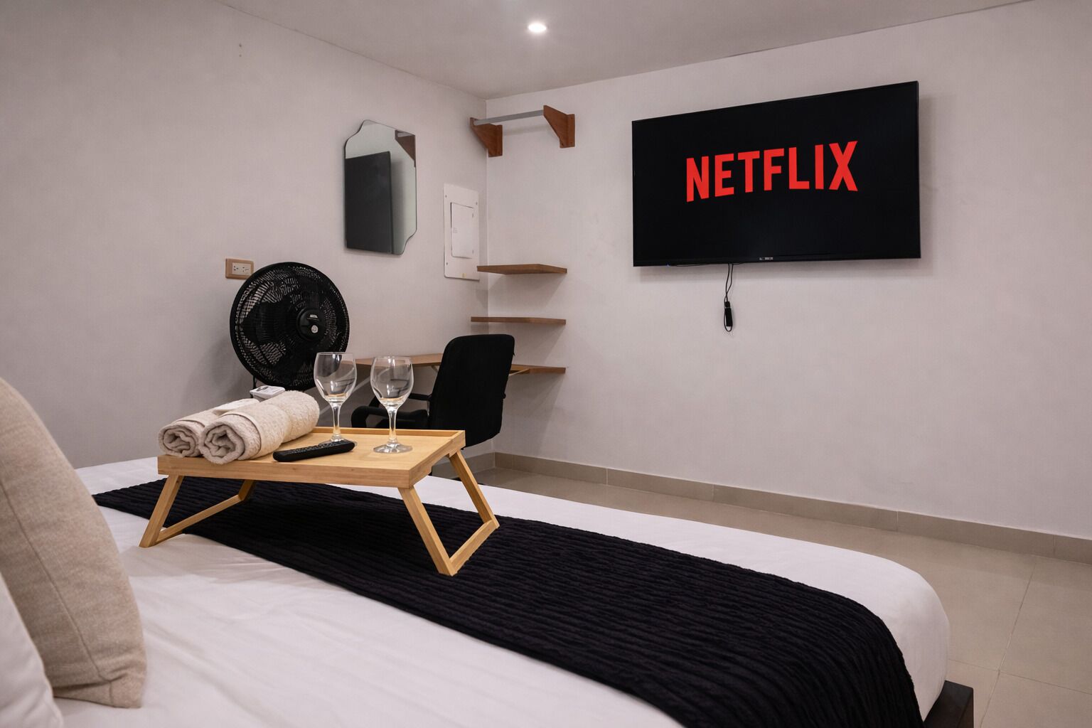 Habitación de hotel con una cama, un televisor montado en la pared, un ventilador, una mesita con un control remoto y vasos, y un estante con un espejo y un enchufe.