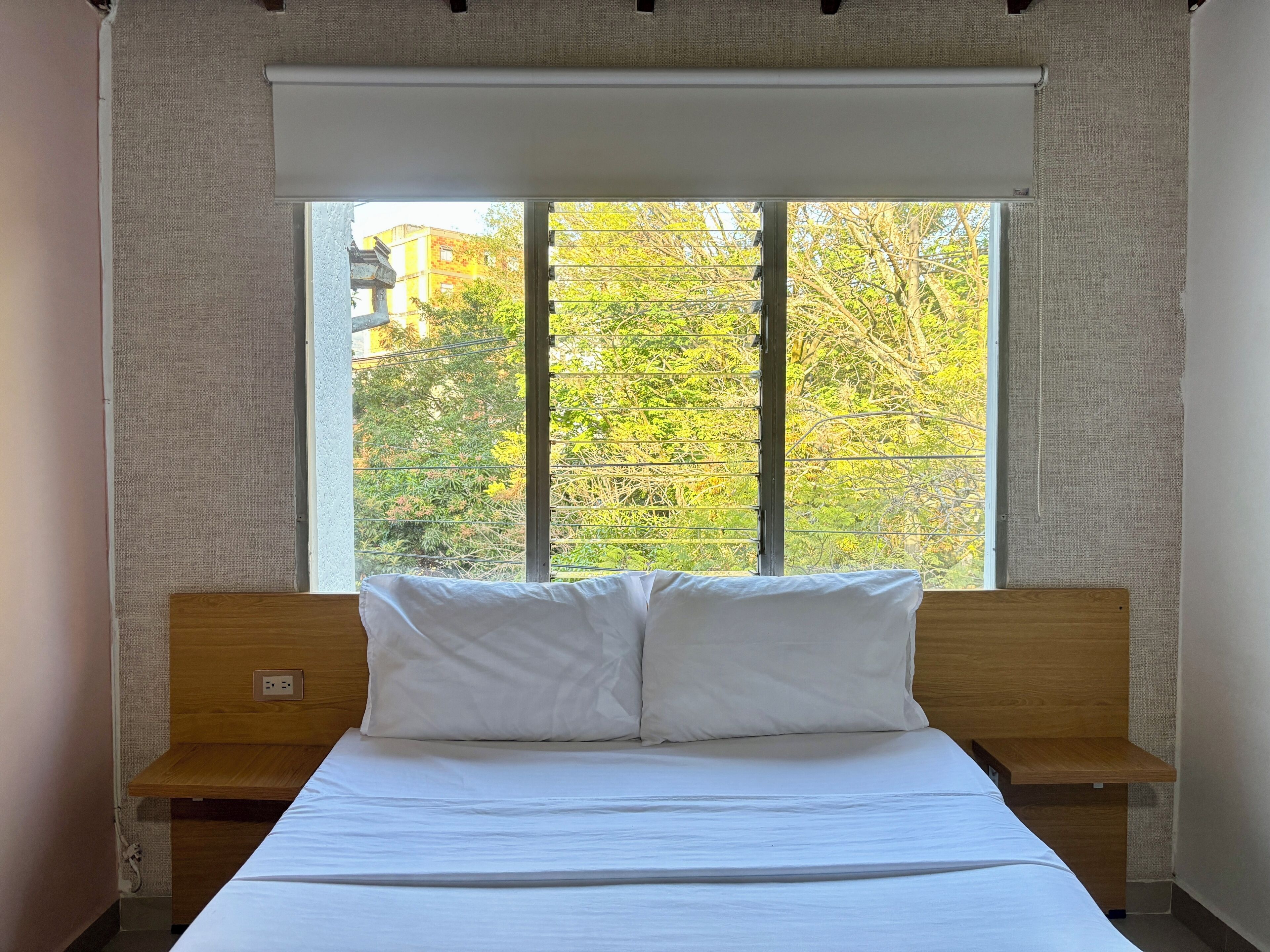 A bedroom with a large window, a wooden headboard, a white bedspread, and a bedside table.