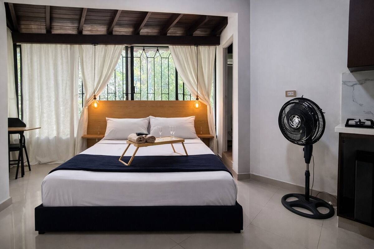 A neatly made bed with a tray of towels and glasses, a standing fan, and a view of greenery through the window.