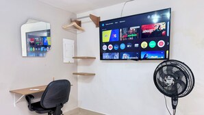 Un espacio de oficina moderno con un televisor montado que muestra diferentes servicios de streaming, un escritorio con una silla y un ventilador de pie.