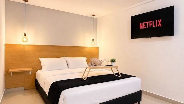 Una habitación de hotel moderna con cabecera de madera, cama negra y blanca, dos lámparas colgantes, una mesita de noche, una bandeja con toallas y vasos, y un televisor mostrando 'NETFLIX'.