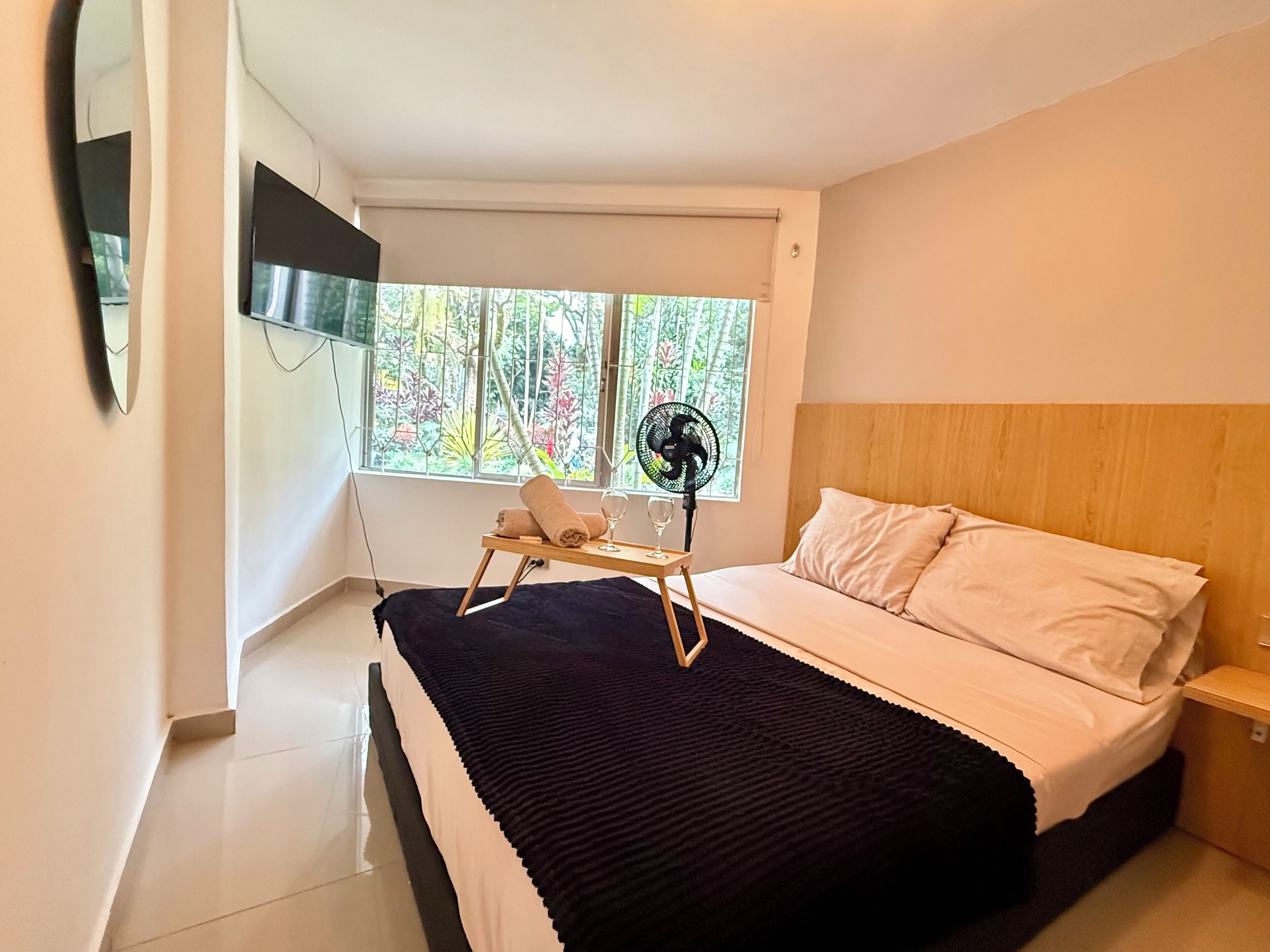 Un dormitorio con cama, mesita de noche, ventilador, ventana con vistas a árboles y televisor montado en la pared.