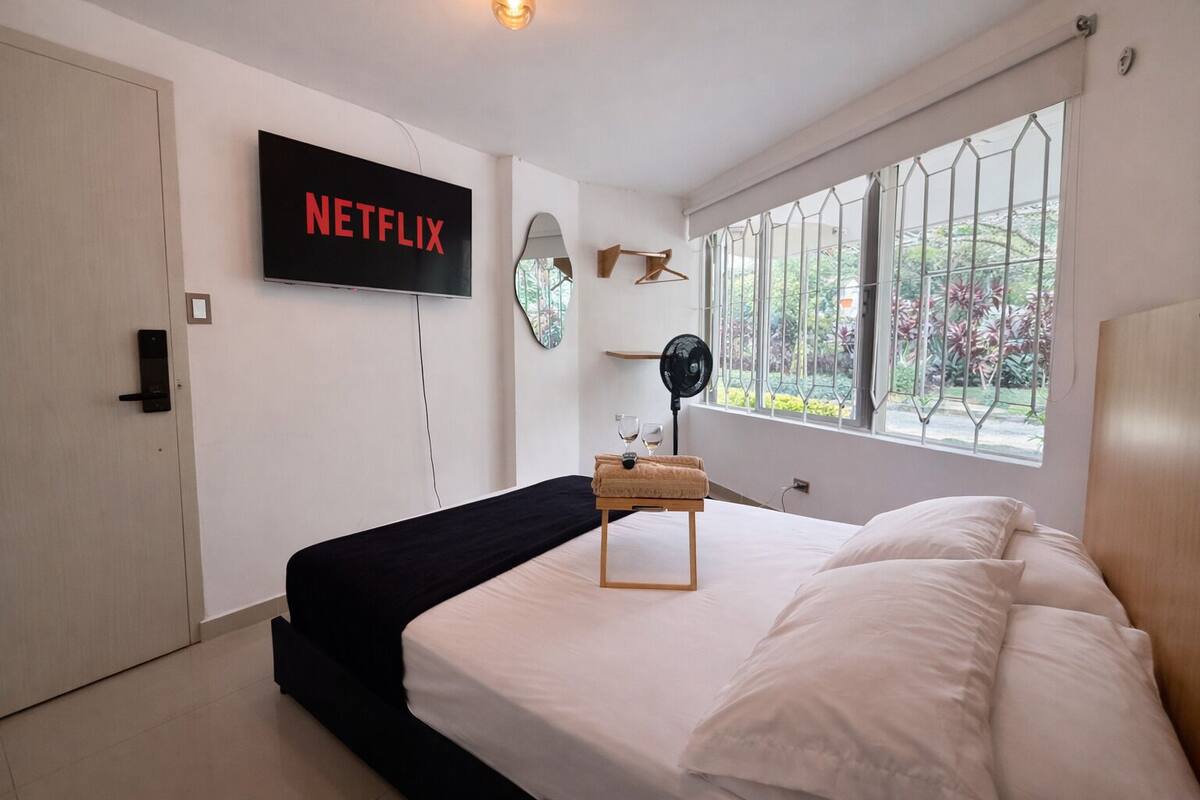A bedroom with a bed, a wall-mounted TV, a window with a view, and a bedside table with a fan.