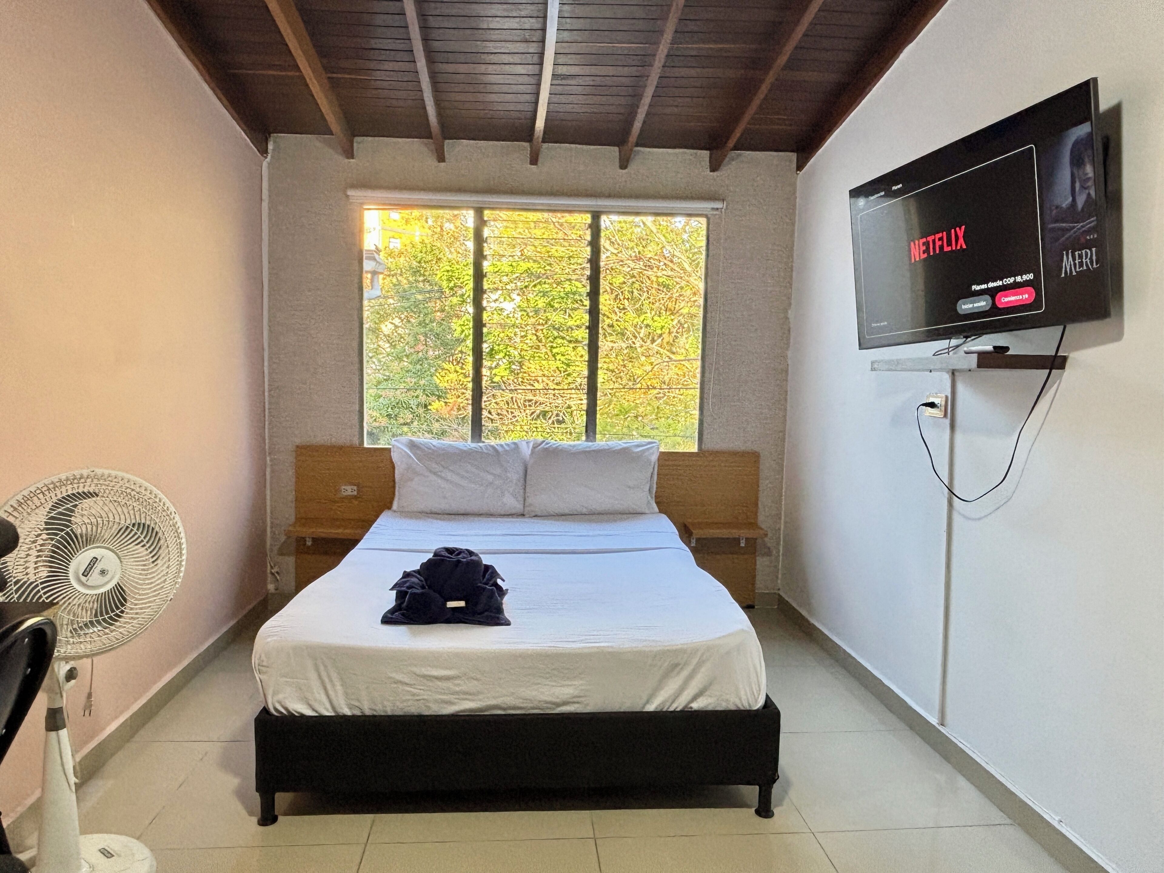 A bedroom with a bed, a TV mounted on the wall, a window, and a fan.