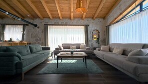 Un salon avec un mur en pierre, des poutres apparentes en bois au plafond et une grande fenêtre.