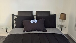 Un lit recouvert d’une couverture sombre, avec deux oreillers, un tableau encadré accroché au mur, une lampe de chevet et une petite table en verre.