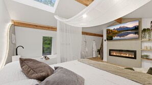 A bedroom with a bed, a fireplace, a large window, and a painting of a mountain landscape.