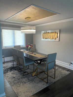 A dining area with a rectangular table, four chairs, a chandelier, a wall-mounted abstract art piece, and a window with blinds. - Poolside Estate (Dubuque)
