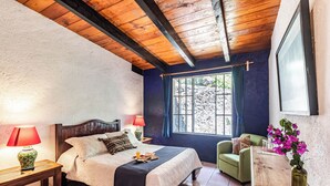 A bedroom with a wooden ceiling, a bed with a headboard, a nightstand, a green armchair, a wooden table, and a window with curtains.