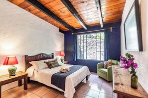 Quarto com teto de madeira, cama com cabeceira, mesa de cabeceira, poltrona verde, mesa de madeira e janela com cortinas.