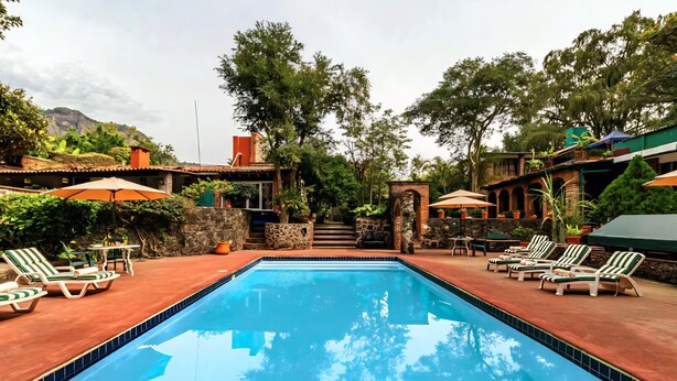 A rectangular swimming pool with lounge chairs and umbrellas on both sides, surrounded by stone walls and greenery.
