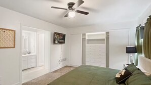 A bedroom with a ceiling fan, a bed with a green bedspread, a TV mounted on the wall, and a walk-in closet.