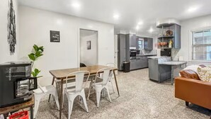 A modern kitchen with grey cabinets, a dining area with a wooden table and white chairs, and a living area with a sofa and a TV.