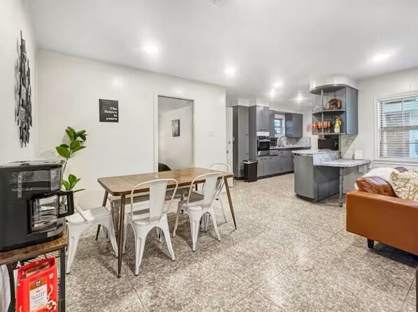 A modern kitchen with grey cabinets, a dining area with a wooden table and white chairs, and a living area with a sofa and a TV.