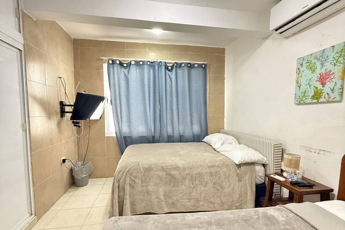 A hotel room with two beds, a shower, a small table with a lamp, and a wall-mounted air conditioner.