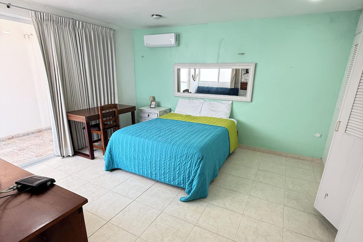A room with a bed, a desk, a chair, a window with curtains, an air conditioning unit, and a mirror on the wall.