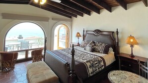 A bedroom with a bed, a nightstand, a lamp, a chair, a small table, and a view through an arched window. - Wonderful 3-bedroom house in Las Gaviotas perfect for your getaway (Las Gaviotas)