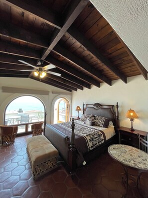 Ein Schlafzimmer mit Bett, Nachttisch, Tisch und Blick durch ein rundbogenförmiges Fenster.