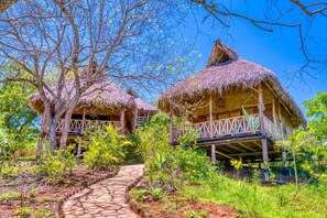 A thatched-roof bungalow with a wooden deck, surrounded by lush greenery and a clear sky. - Eden Yoga & Wellness Resort (San Juan del Sur)