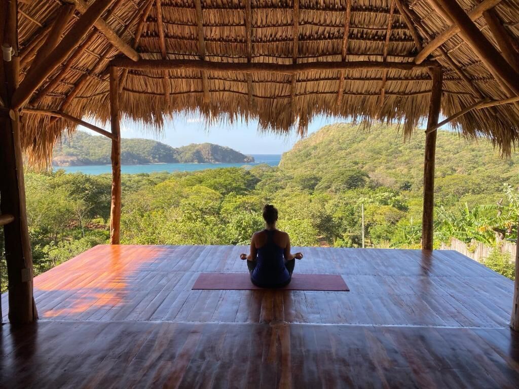 Una persona meditando en una terraza de madera con techo de paja, con vistas a un paisaje exuberante y un cuerpo de agua.