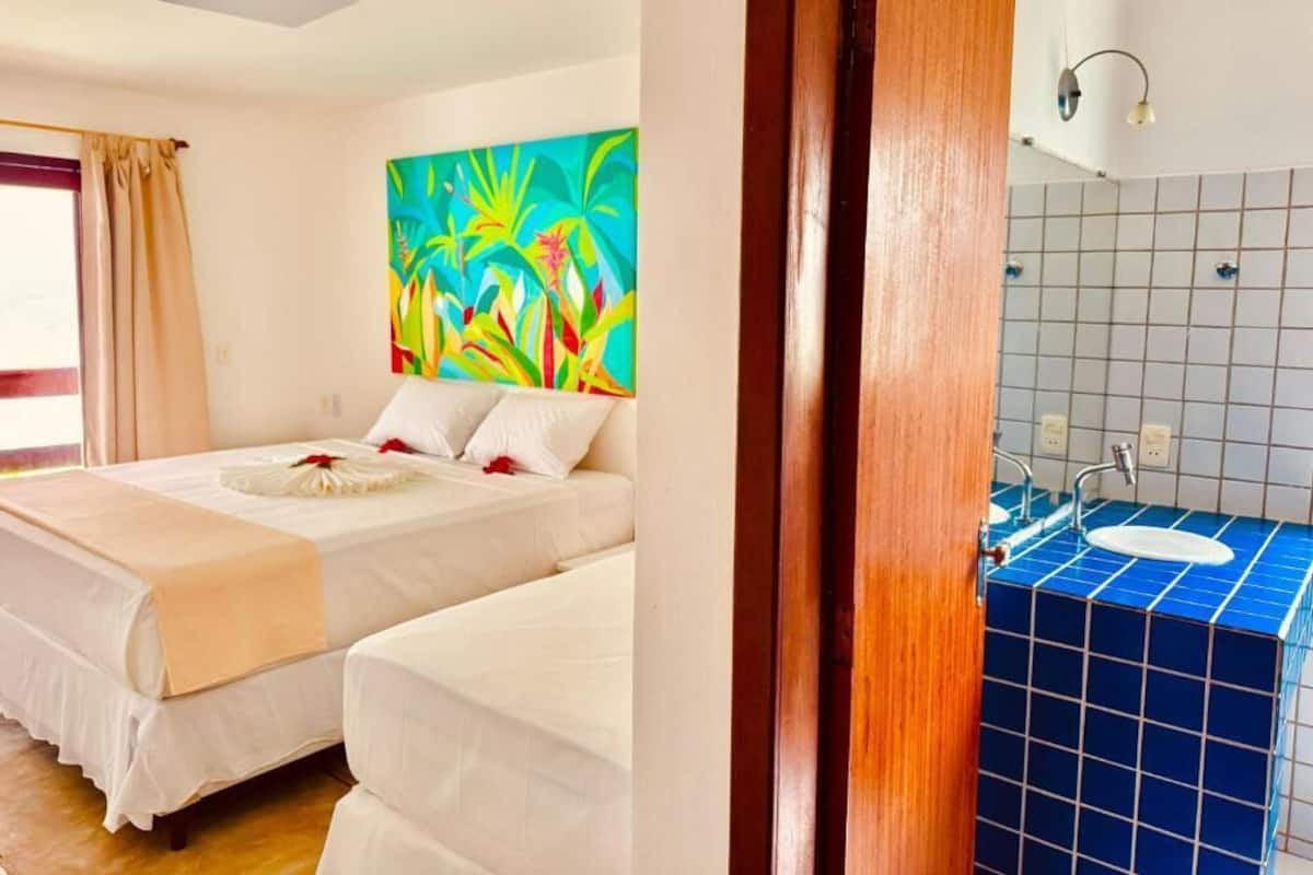 A hotel room with two beds, a colorful painting, a bathroom with blue tiles, and a wooden door.