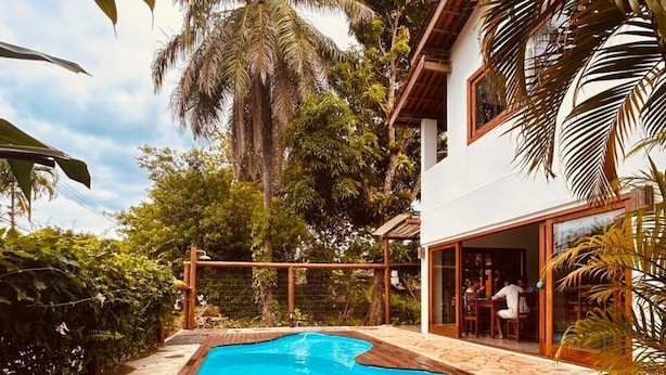 Una piscina con muelle de madera, rodeada de exuberante vegetación tropical y una casa con amplios ventanales.