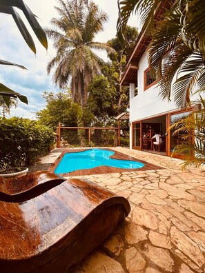 Una piscina con muelle de madera, rodeada de exuberante vegetación tropical y una casa con amplios ventanales.