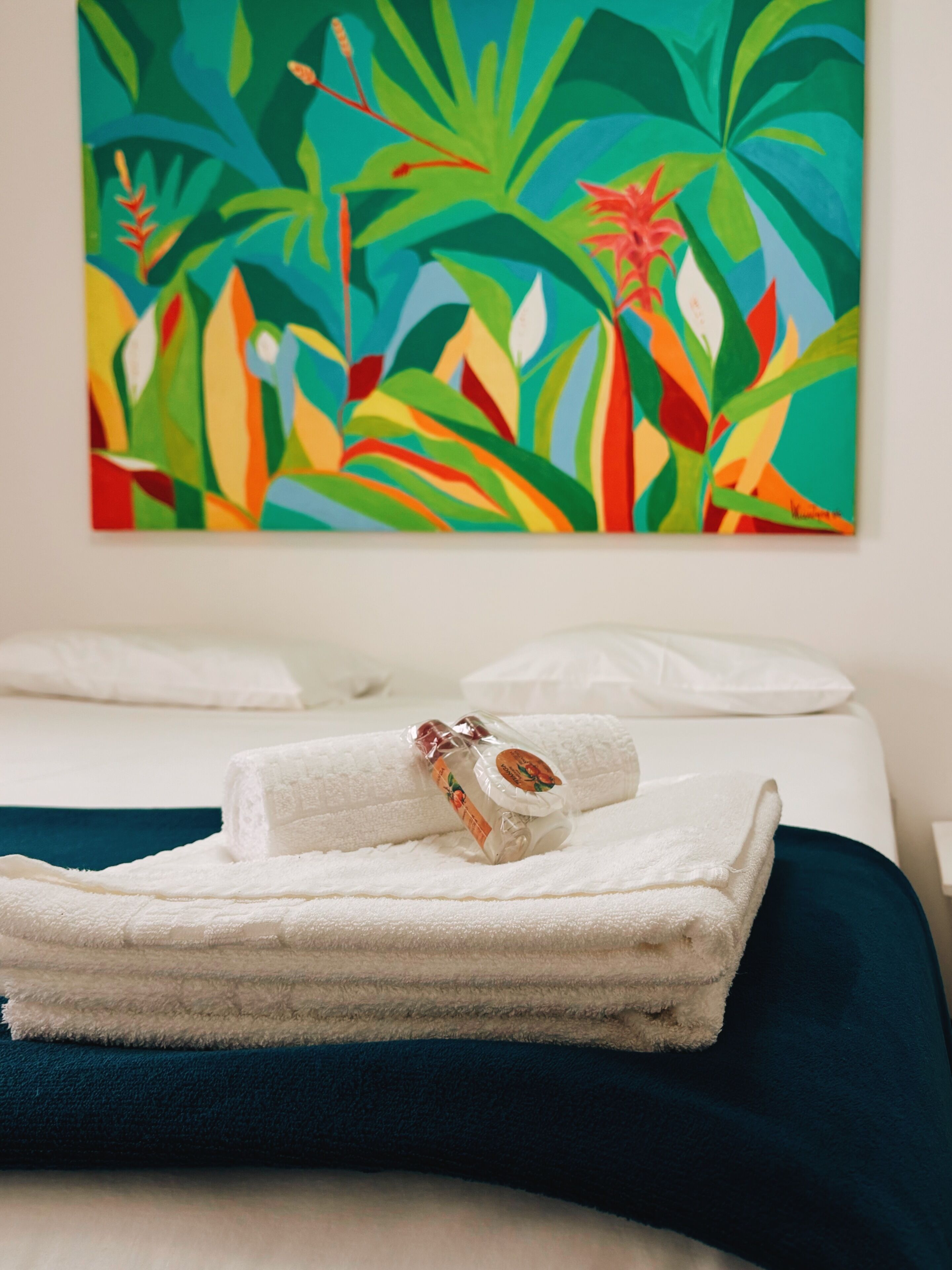 Een hotelkamer met een bed, een keurig opgevouwen witte handdoek en een kleurrijk schilderij met een tropisch thema aan de muur.