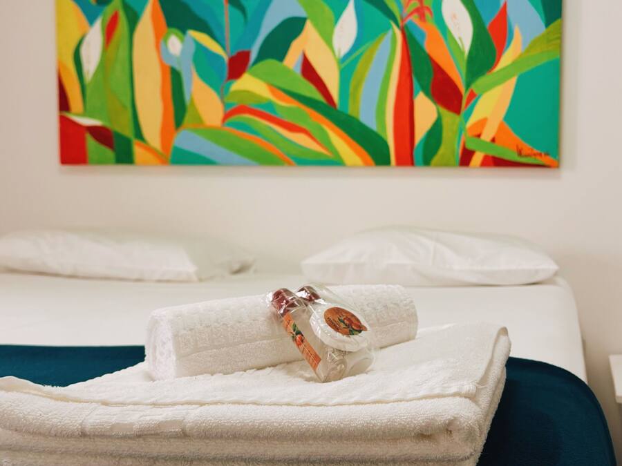 Een hotelkamer met een bed, een keurig opgevouwen witte handdoek en een kleurrijk schilderij met een tropisch thema aan de muur.