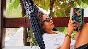 Una persona descansando en una hamaca junto a una ventana, con un libro, rodeada de abundante follaje verde.