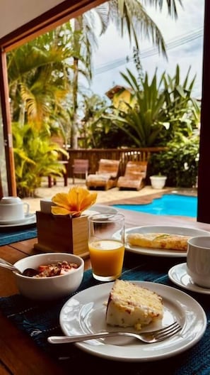 A dining table with breakfast items, a glass of orange juice, and a slice of cake, overlooking a pool and tropical plants.